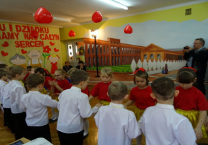 Dzieci z gr. IV: dziewczynki w czerwonych podkoszulkach i żółtych spódniczkach oraz chłopcy w białych koszulach i czarnych spodniach tańczą w parach dla Dziadków.